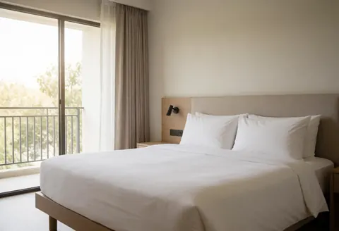 Minimalist hotel room featuring a large bed, curtains, and a view of trees from the balcony.