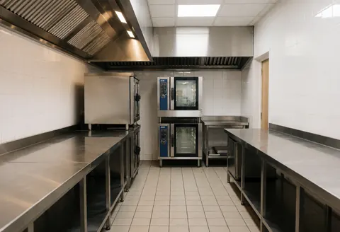 Empty professional kitchen with stainless steel counters, tiled walls and floor, modern pots and ovens, and bright clean lighting.