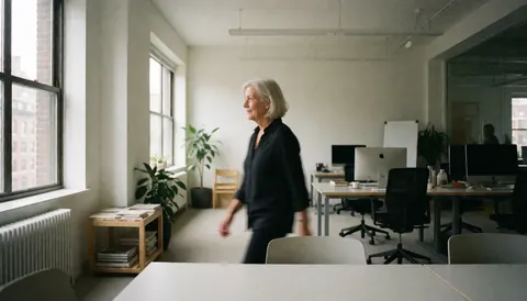 A female architect walks through a bright modern office with desks, chairs, and plants, ideal for architecture and interior design services.