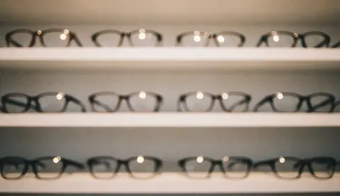 Dark framed eyeglasses displayed on white shelves against a blurred background with warm lights, suitable for commercial text overlays