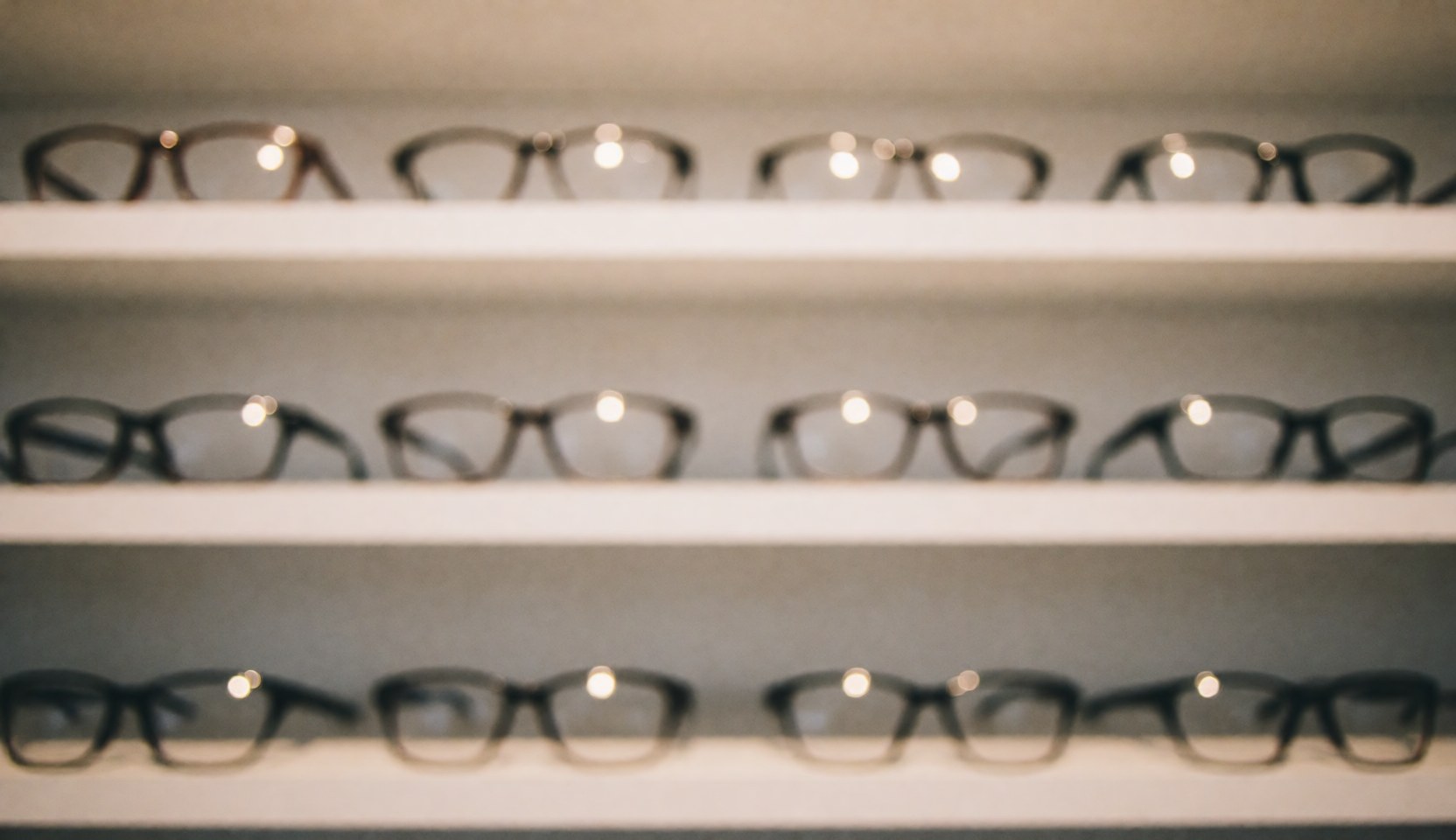 Dark framed eyeglasses displayed on white shelves against a blurred background with warm lights, suitable for commercial text overlays