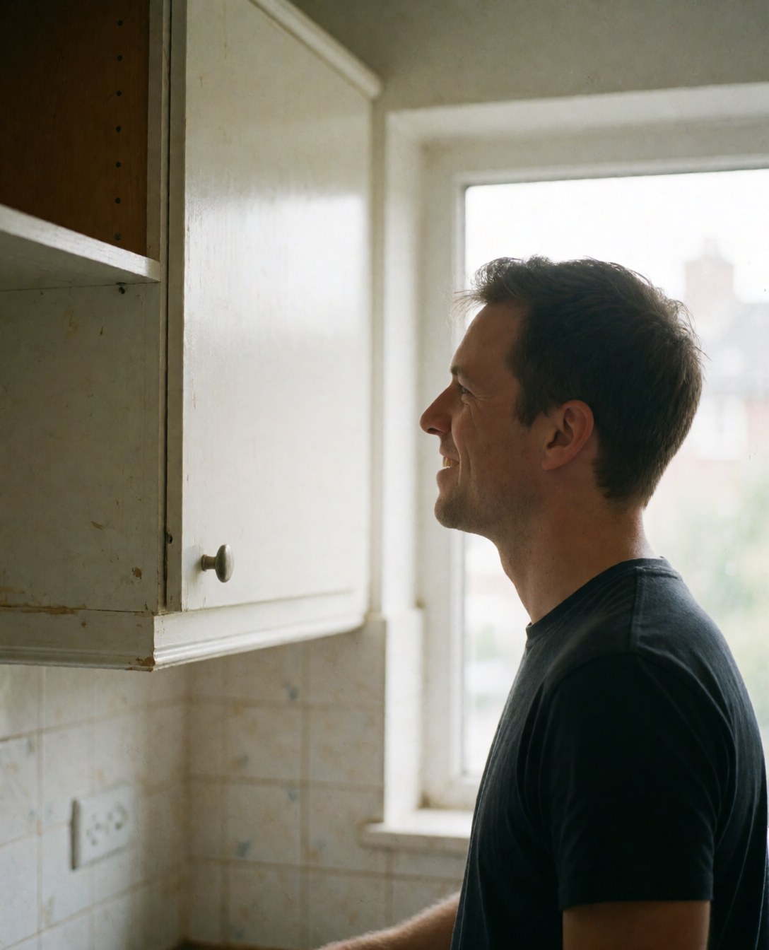 Smiling customer looks at a newly installed kitchen cabinet with natural light from a window and tiled walls