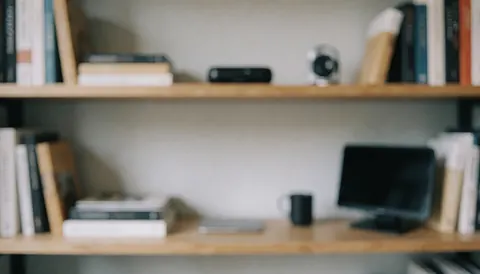 Wooden bookshelf holding books, a laptop, a smartphone, and tech gadgets against a soft blurred background suitable for text overlay
