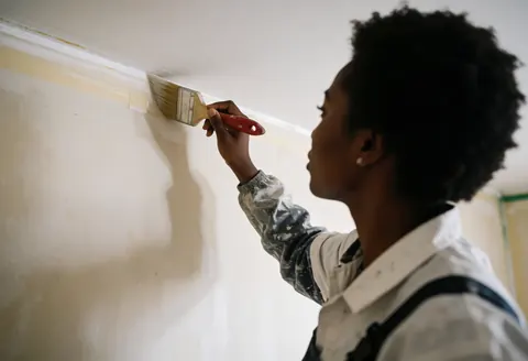 A professional female painter uses a brush to apply paint to a wall, demonstrating precision and technique in her interior painting work.