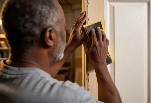 A man sanding a wooden door frame with sandpaper in a well-lit workshop.