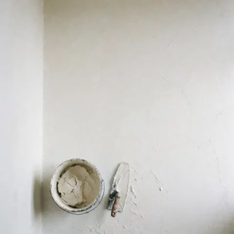 A newly plastered white wall with a paint bucket and roller, ideal for renovation or therapy office decor with soft natural lighting