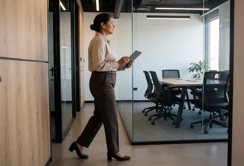 A businesswoman walks down an office corridor holding a tablet while looking toward a meeting room with tables and chairs.