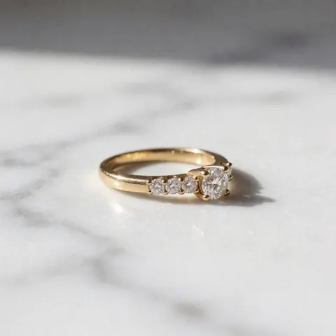 Gold ring with bright diamonds on white marble surface, soft side lighting highlighting the sparkle of the stones
