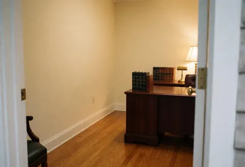 View from the doorway of an empty law office featuring a wooden desk, books, a lamp, cream walls, and polished wood flooring