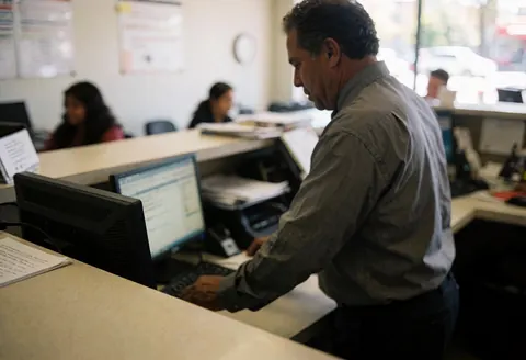 Professional man helping a customer at a computer in a busy office with colleagues in the background