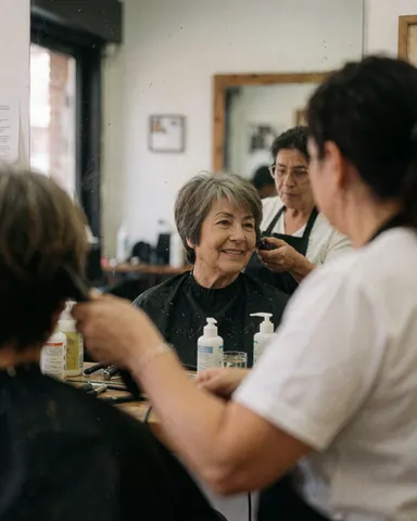 Smiling older woman looks at her new hairstyle in the mirror while a stylist works nearby.