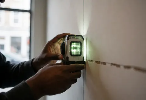 Close-up of a worker's hand using a laser level to mark a straight line on a white wall during interior construction