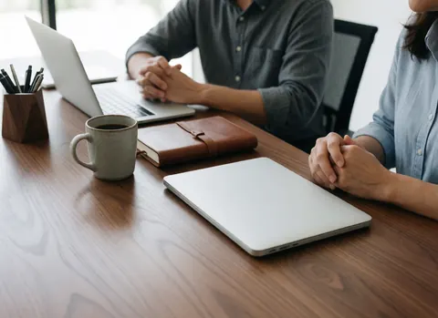 Two people working at a wooden table with a laptop, coffee cup, notebook, and pen holder, illuminated by natural light