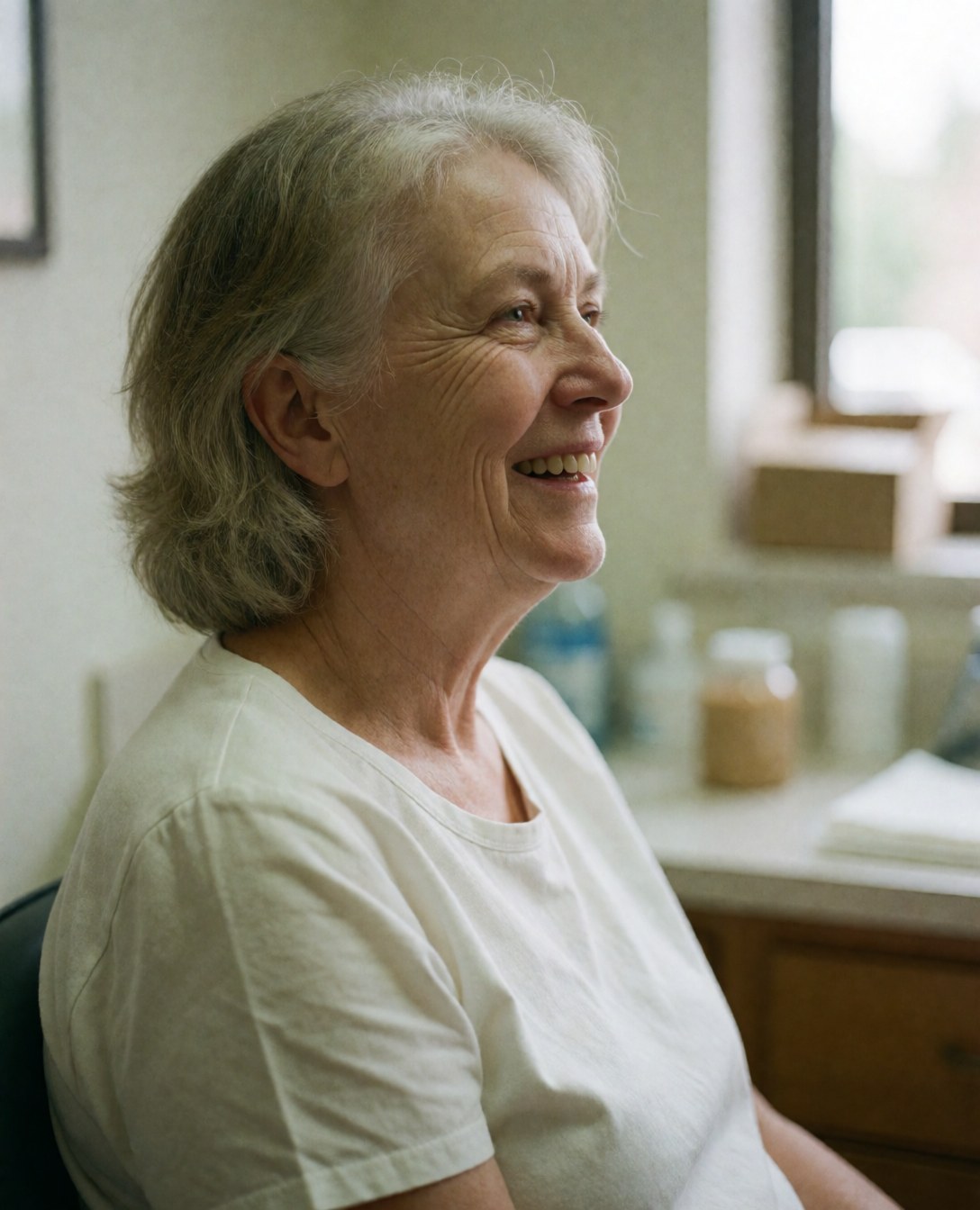 A smiling older woman sits in a dental chair with natural light and medical office background