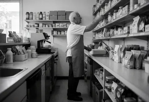 An older man wearing an apron reaches for a product on a store shelf with natural light and visible items in the background
