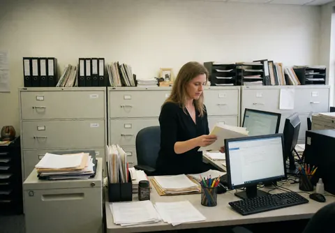 A businesswoman reviews documents at her desk surrounded by file cabinets, computers, and organized papers in a professional setting.