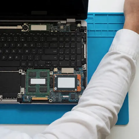 Technician in a white shirt repairing an open laptop on a blue surface.