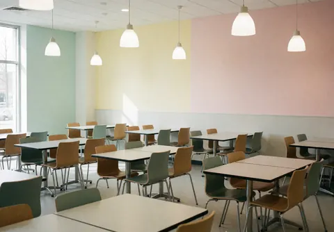Modern university cafeteria featuring empty tables and chairs, pastel walls, hanging lights, and a clean cozy atmosphere for students