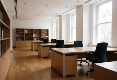 Spacious modern workspace featuring wooden desks, black chairs, bookshelves, and natural light from tall windows