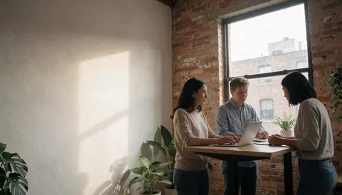 Three people working together at a desk in a coworking space with laptop, brick wall and green plants.