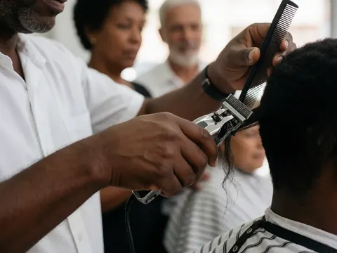 A barber uses electric clippers and a comb to cut a client's hair in a busy barbershop with other customers in the background.