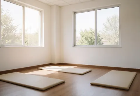 Empty physical therapy room with mats on wood floor, large windows with natural light and white walls