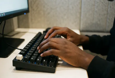 Hands typing on a black keyboard with blue backlight, in front of a computer on a white desk.