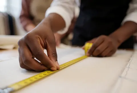 Close up of a seamstress hand measuring white fabric with a yellow tape measure in a sewing workshop with blurred background