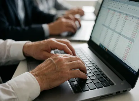 Close-up of adult hands typing on a laptop keyboard with a financial spreadsheet visible on the screen in a professional office setting