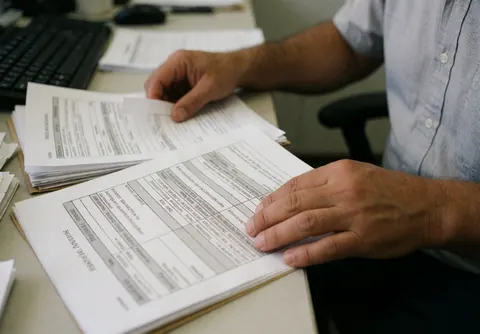 A man in a blue shirt reviews financial papers on an office desk with a keyboard and mouse.