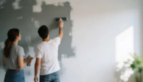 A couple paints a wall with a roller and brush in a room with natural light.