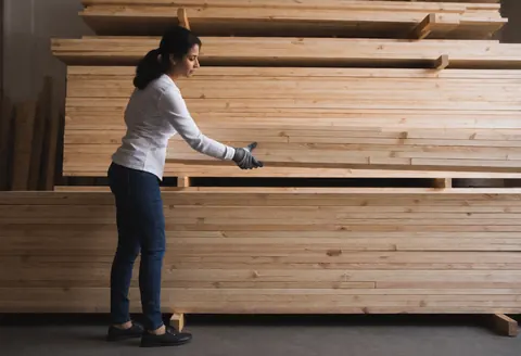 A woman wearing gloves organizes piles of lumber in a hardware store warehouse with soft lighting and a professional atmosphere