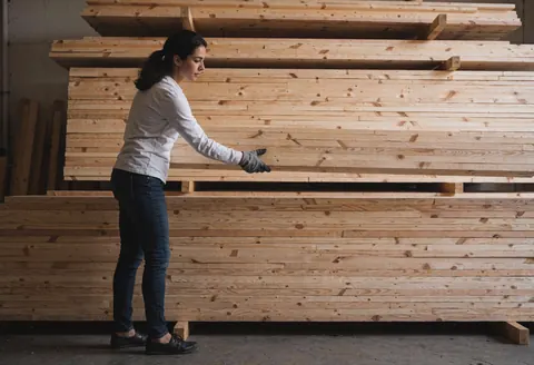 A woman wearing gloves organizes piles of lumber in a hardware store warehouse with soft lighting and a professional atmosphere