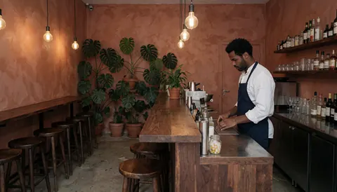 Interior view of a cocktail bar featuring a wooden counter, potted plants, chairs, and a bartender preparing drinks