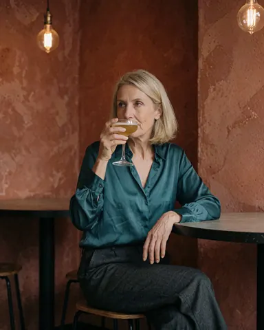 A mature woman relaxes with a cocktail at a bar featuring terracotta walls and Edison bulbs.