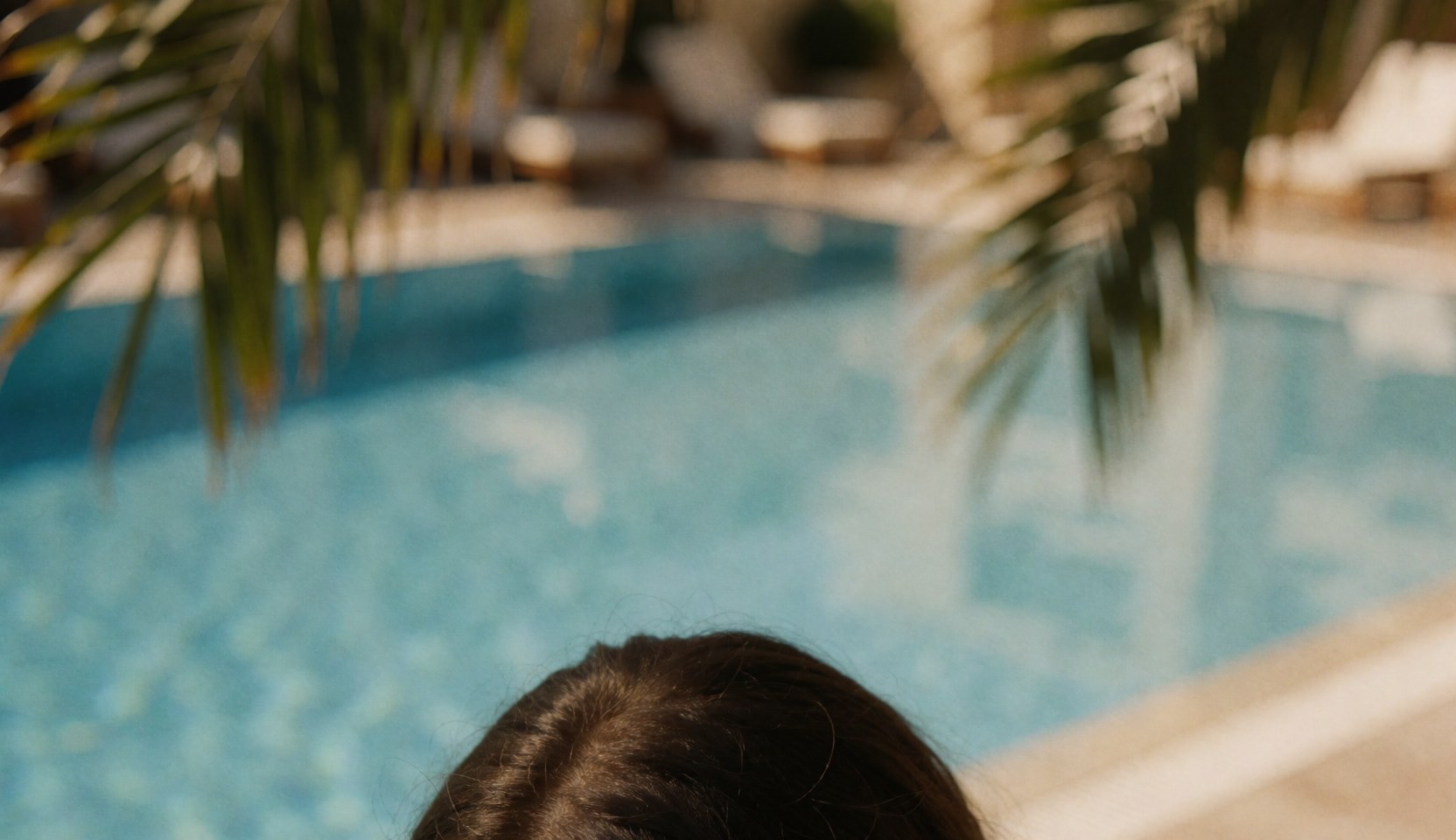 A person looks at a blue pool with palm leaves in foreground, in a relaxed and sunny setting.