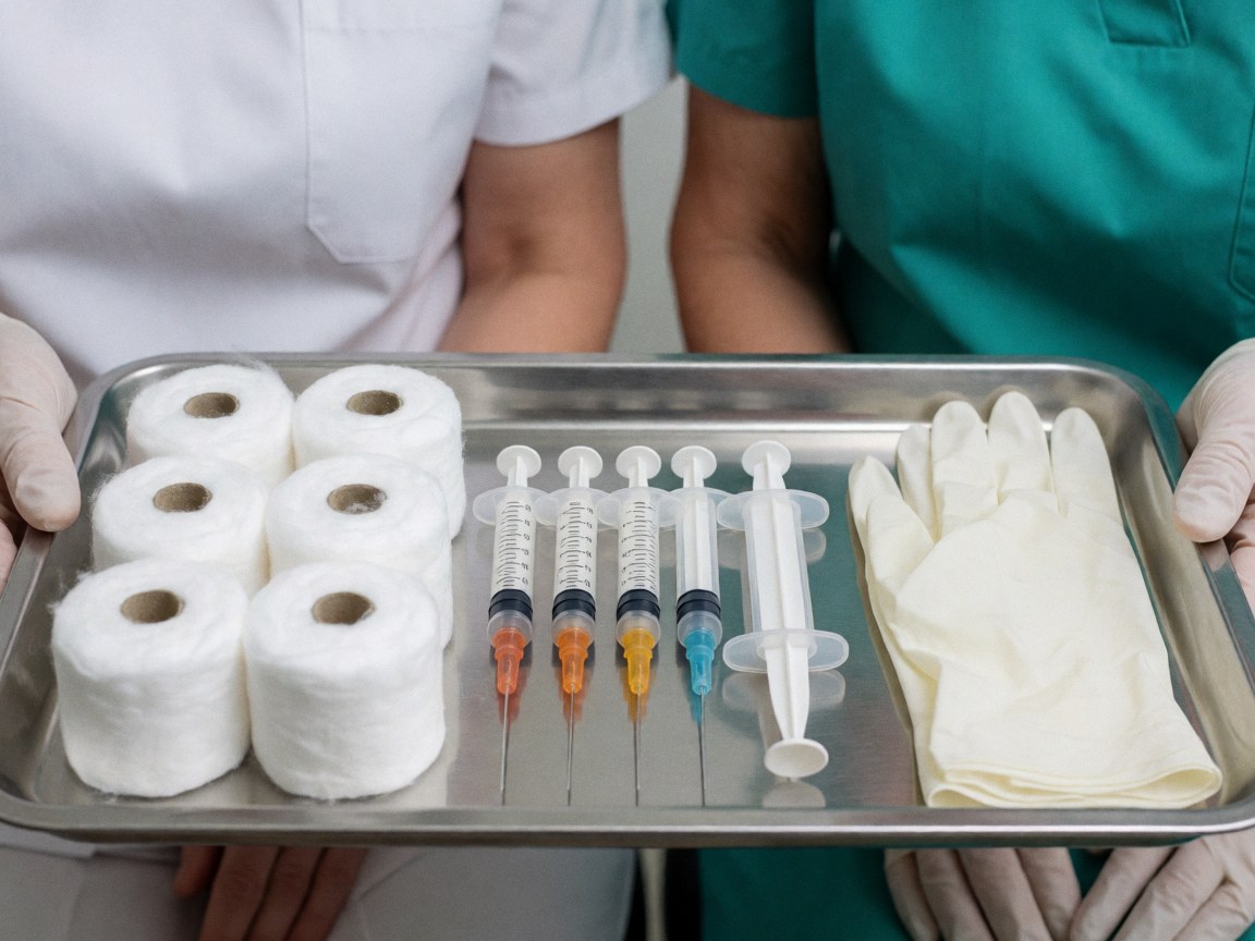 Two medical professionals hold a metal tray with syringes, gloves, and cotton rolls for clinical procedures.