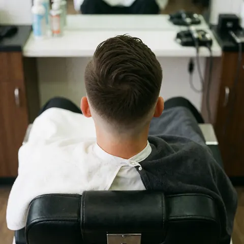 Close-up of a man receiving a fade haircut in a barber chair with visible towels and grooming products under bright lighting