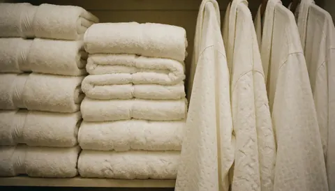 Neatly folded white towels and hanging white robes in a hotel or spa closet, ready for use.