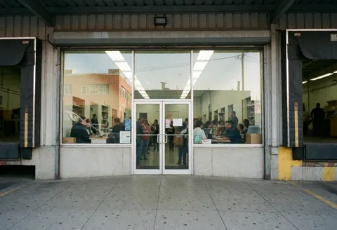 Industrial factory exterior featuring a large window, metal facade, glass doors, and employees working inside the building.