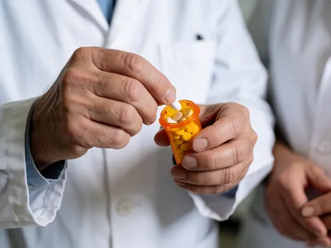 An adult pharmacist in a white coat counts pills in an orange bottle with a blurred background and professional lighting.