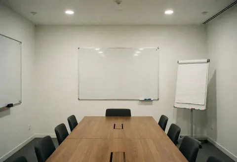 Modern meeting room with wooden table, black chairs, whiteboards, and white walls.