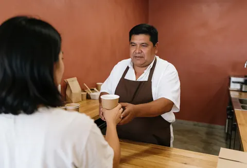 A man serves a drink in a paper cup to a customer in a cafe with terracotta walls and a wooden counter.