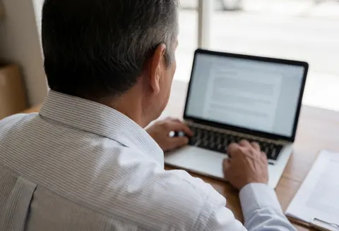 Back view of an older man typing on a laptop near a window with natural light, focused on remote work tasks.