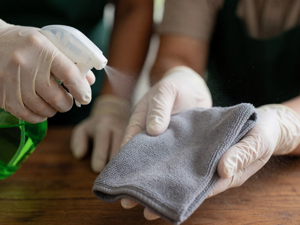 Close-up of a gloved hand spraying cleaning solution onto a gray cloth on a wooden table