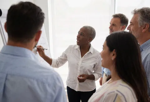 A consultant explains charts on a whiteboard while a group of people attentively watches in an office meeting.