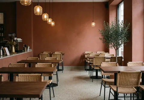 Spacious empty cafe interior featuring terracotta walls, wooden tables, rattan chairs, warm lighting, and a potted plant in the corner