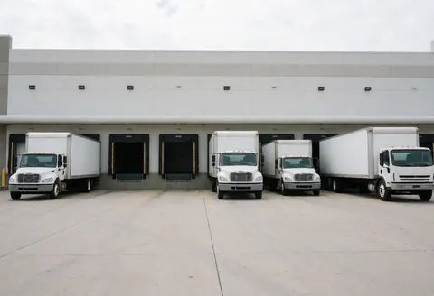 Modern warehouse exterior with several cargo trucks parked in front of loading docks on a concrete floor under a cloudy sky.