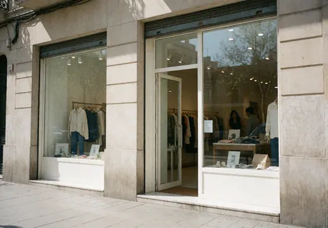 Modern fashion boutique featuring an open entrance, clean display window, and stone facade with visible clothing racks inside.
