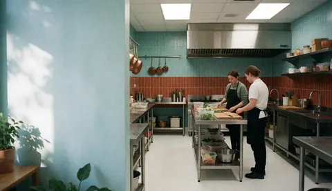 A modern catering kitchen featuring turquoise walls, red tiles, stainless steel tables, and two chefs preparing food.